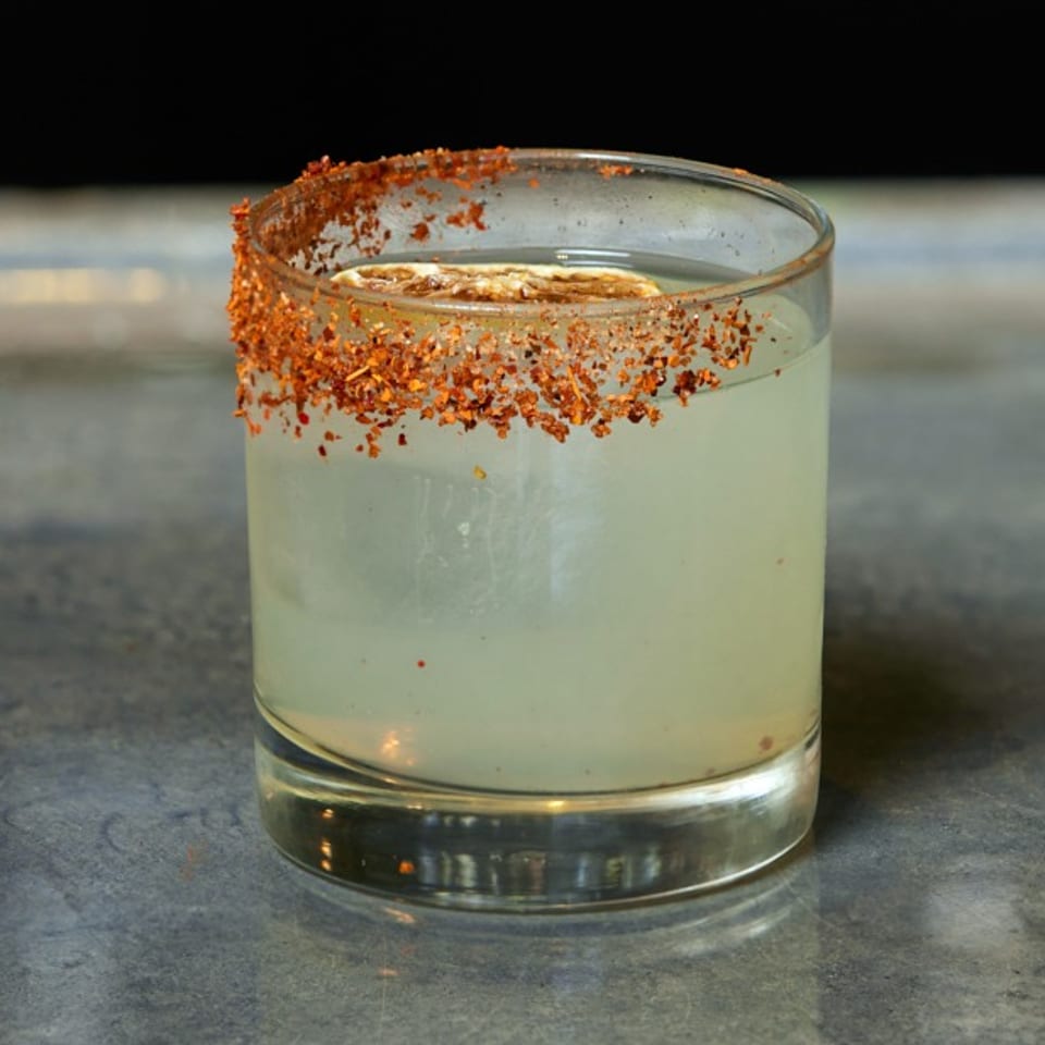 A light-colored cocktail in a glass with a chili-salt rim and a lime slice.