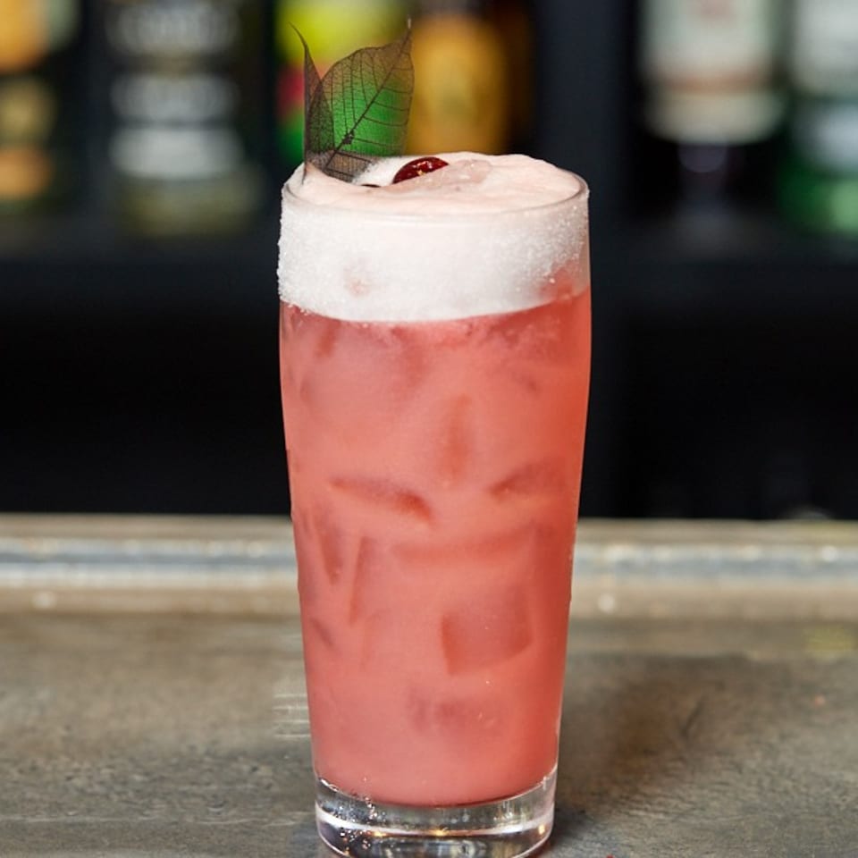 Pink cocktail with ice, foamy top, sugar rim, and a leaf garnish on a bar counter.