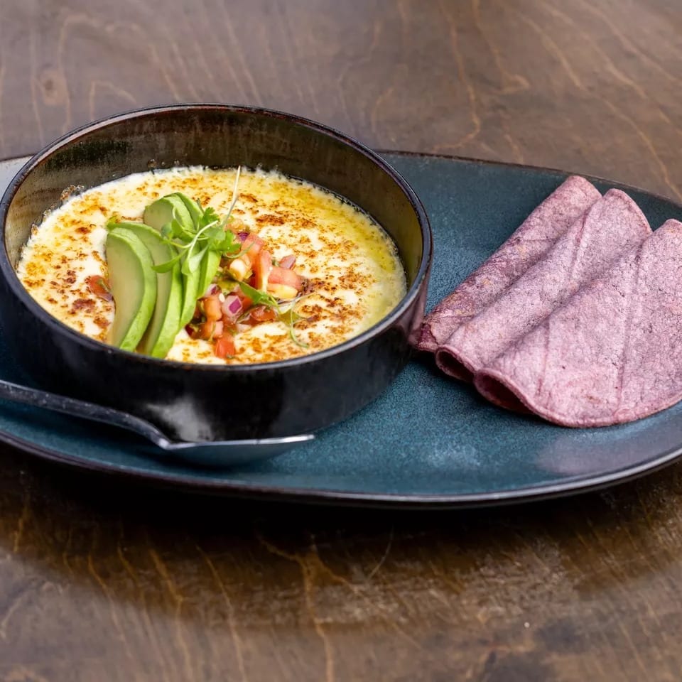 Melted cheese dip with avocado, pico de gallo, and purple tortillas on a dark plate.