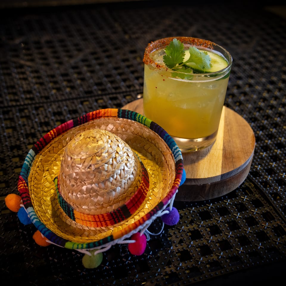 A cucumber margarita with a chili-salt rim next to a miniature sombrero on a dark textured surface.