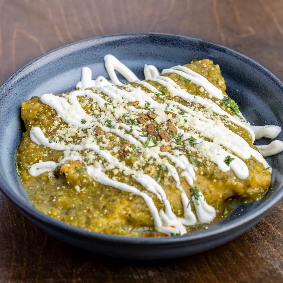 Green enchiladas topped with crema, cotija cheese, and pumpkin seeds in a dark blue bowl.