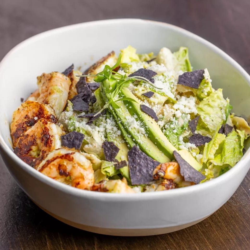 Grilled shrimp salad with avocado, blue tortilla chips, and crumbled cheese in a white bowl.