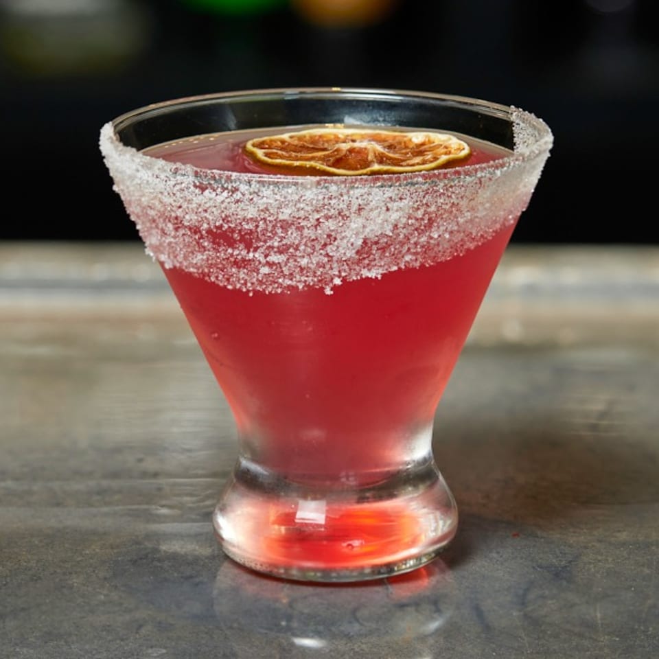 A vibrant red cocktail in a sugar-rimmed martini glass, garnished with a dried orange slice.