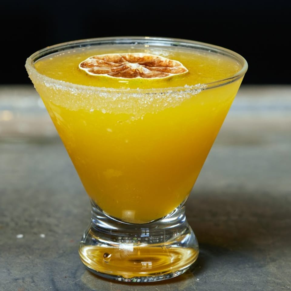 A vibrant yellow cocktail in a martini glass, garnished with a dried citrus slice and sugar rim.