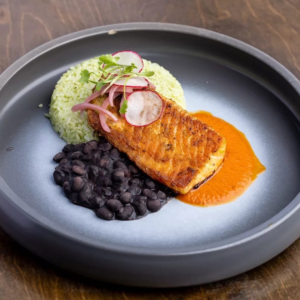 Pan-seared fish with green rice, black beans, and red sauce, garnished with radishes and herbs.